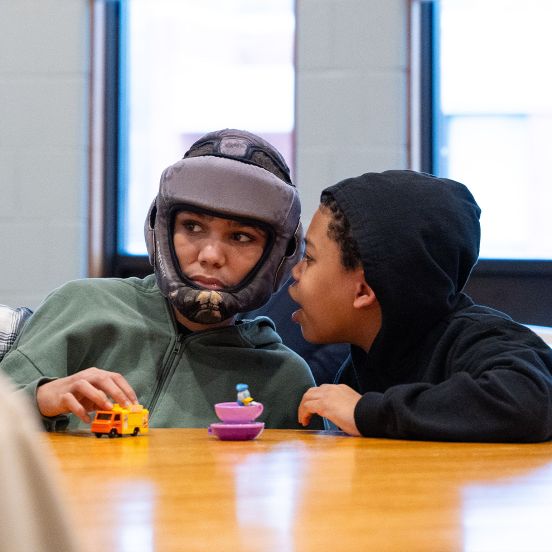 Two young boys sitting next to each other while one on the right is whispering into the ear of the one on the left